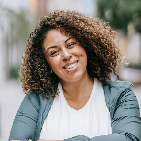 Au&szlig;enportrait einer jungen Frau mit braunen locken, die mit verschr&auml;nkten Armen freundlich in die Kamera l&auml;chelt.