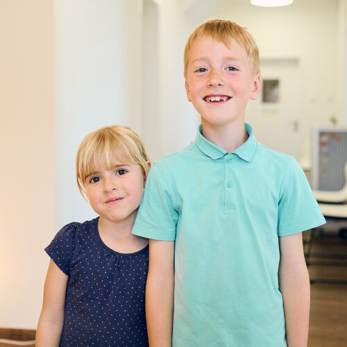 Zwei l&auml;chelnde Kinder, ein M&auml;dchen und ein Junge mit Zahnl&uuml;cke, stehen in einem hellen Flur einer Zahnarztpraxis.