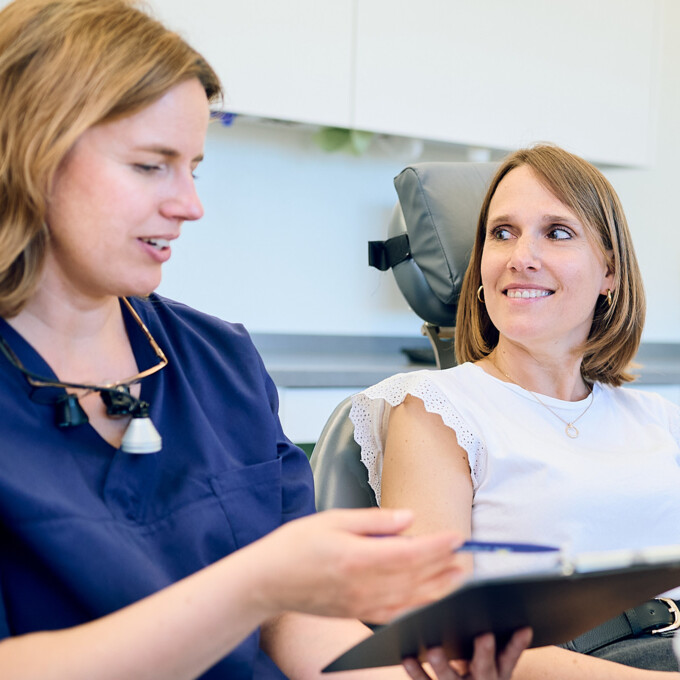 Zahn&auml;rztin Anna Rothe erl&auml;utert einer Patientin auf dem Behandlungsstuhl Details anhand einer Klemmmappe.
