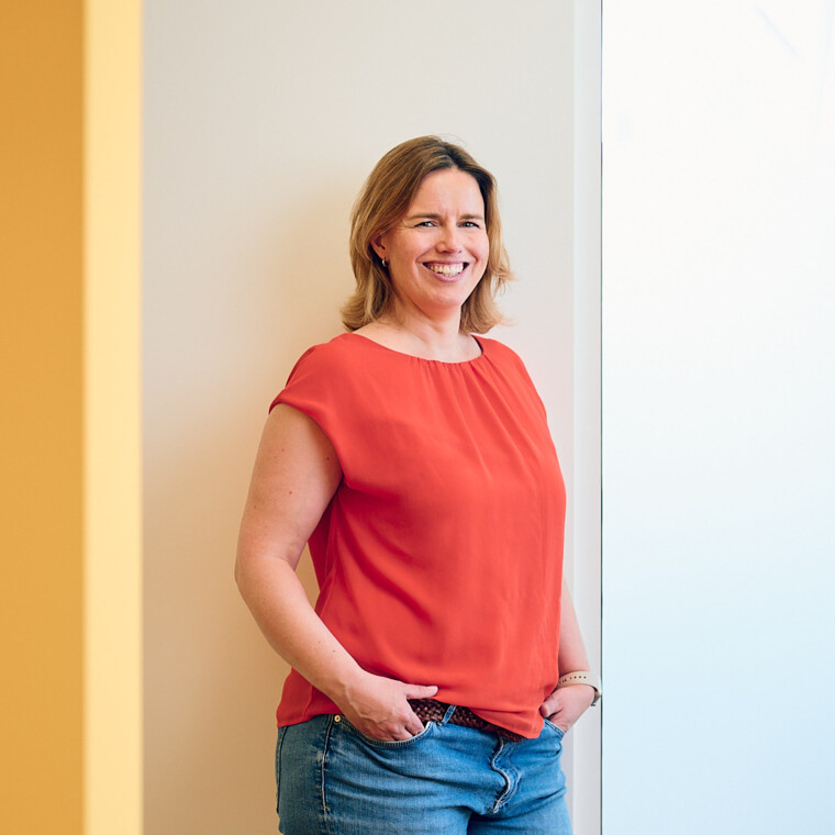 Zahn&auml;rztin Anna Rothe steht in orangener Bluse in Ihrer Praxis und l&auml;chelt sympathisch in die Kamera