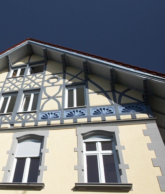 Fassade der Praxis Breisach mit blauem Zierfachwerk und hellgelben W&auml;nden unter tiefblauem Himmel.