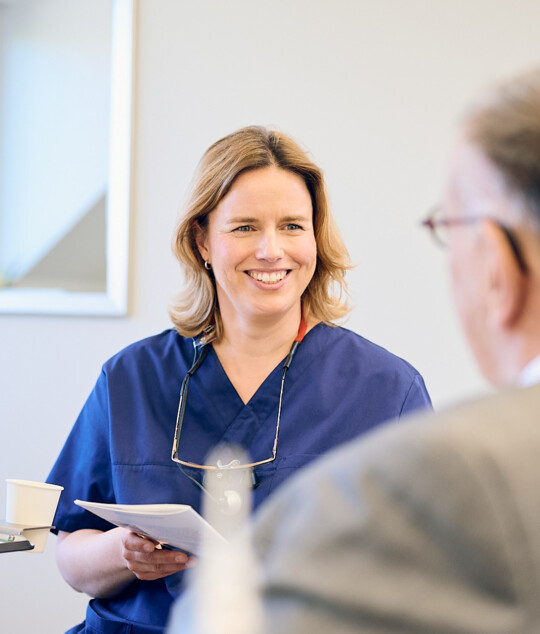 Zahn&auml;rztin Anna Rothe im Beratungsgespr&auml;ch mit einem Patienten, l&auml;chelnd mit Informationsbrosch&uuml;re in der Hand.