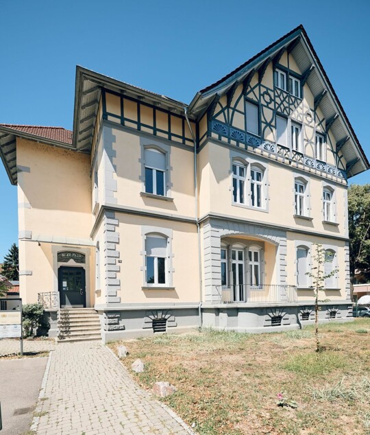 Fassade der Praxis Breisach mit blauem Zierfachwerk und hellgelben W&auml;nden unter tiefblauem Himmel.