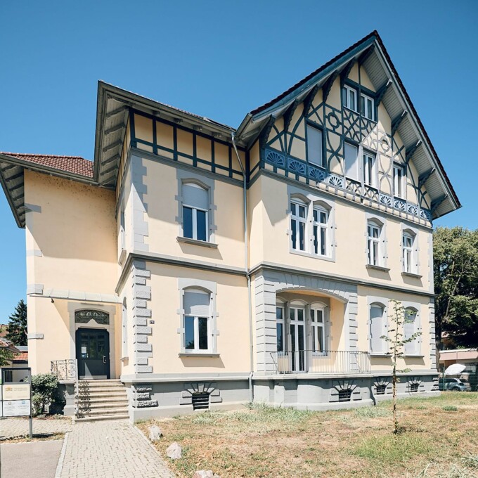 Fassade der Praxis Breisach mit blauem Zierfachwerk und hellgelben W&auml;nden unter tiefblauem Himmel.