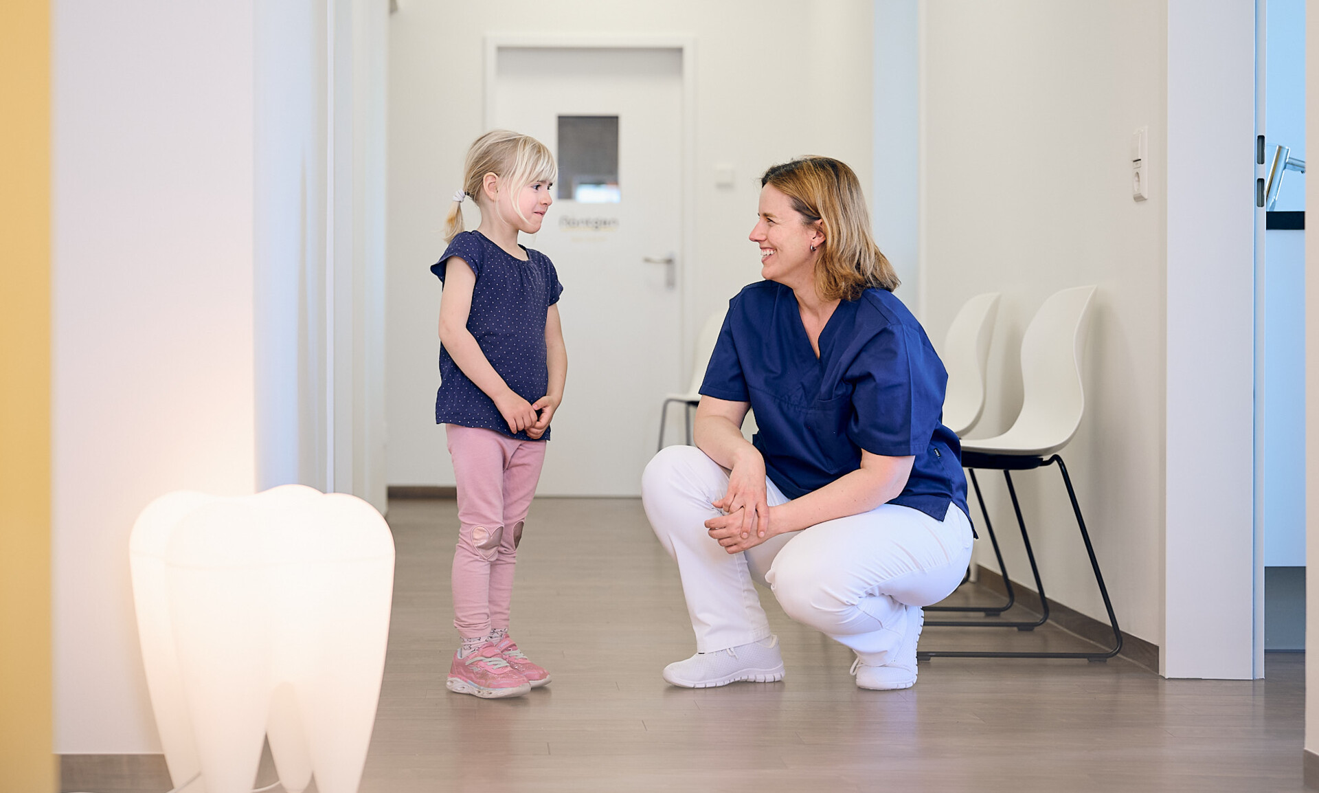 Zahn&auml;rztin Anna Rothe spricht auf Augenh&ouml;he mit einem kleinen M&auml;dchen im hellen Flur ihrer modernen Praxis in Breisach.