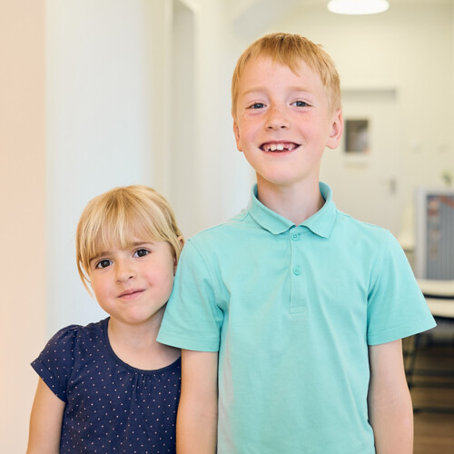 Zwei l&auml;chelnde Kinder, ein M&auml;dchen und ein Junge mit Zahnl&uuml;cke, stehen in einem hellen Flur einer Zahnarztpraxis.